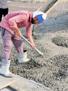 Worker in a pink shirt spreading concrete on a construction site with a shovel.